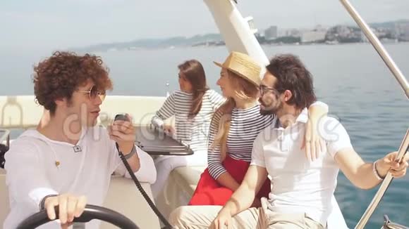 年轻的卷发男子驾驶着一艘海船与他快乐的朋友男人和女孩坐在一起享受着视频的预览图