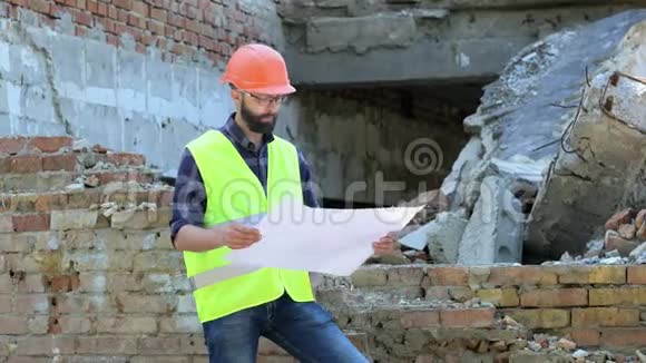 漂亮的胡子建设者在橙色头盔上被摧毁的建筑背景认真的建筑工人分析图纸视频的预览图