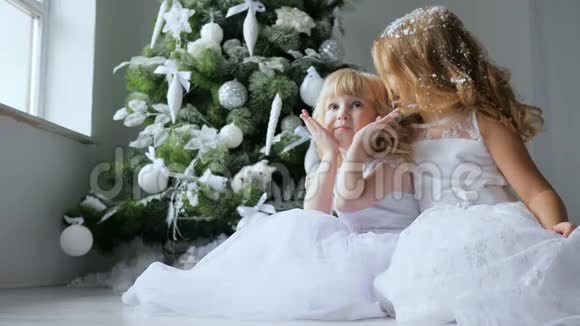 寒假穿着白色连衣裙的姐妹们在新年前夕在装饰的杉树旁边的地板上拍照视频的预览图