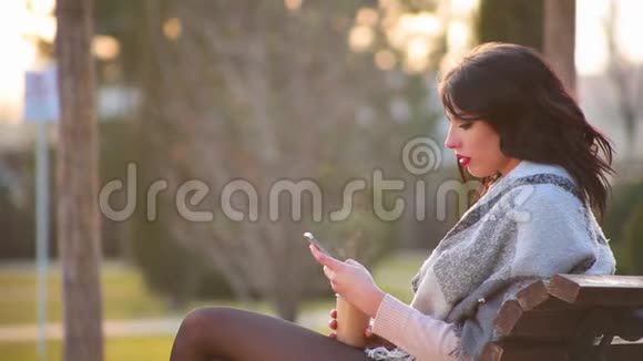 漂亮可爱的黑发女孩坐在城市公园的长凳上喝热咖啡里面有蒸汽和写在电话里视频的预览图