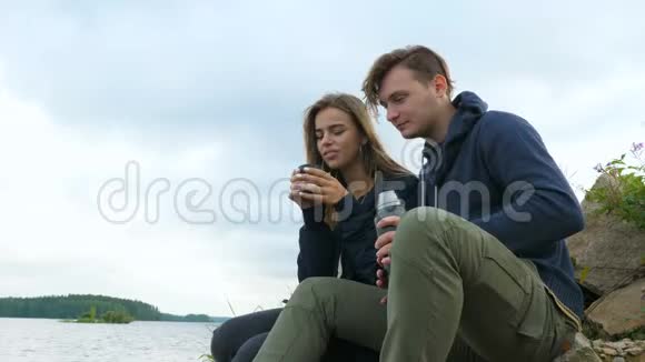 一个男人和一个女孩坐在河岸上喝热水瓶里的茶视频的预览图