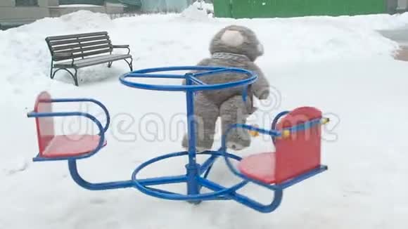 冬天下雪时孤独的泰迪熊在操场上的红色蓝色旋转木马上旋转孤独概念视频的预览图