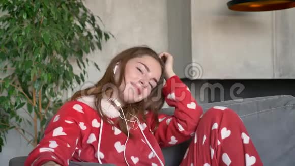 漂亮的年轻女子穿着红色睡衣通过耳机跳舞和听音乐坐在客厅的沙发上视频的预览图