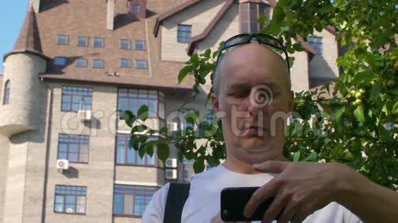 光头男用树和建筑物在城市智能手机上编辑视频视频的预览图