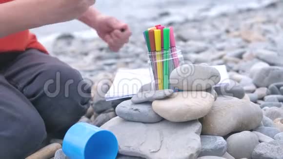 特写小男孩坐在海边的石滩上手里拿着记号笔作画视频的预览图
