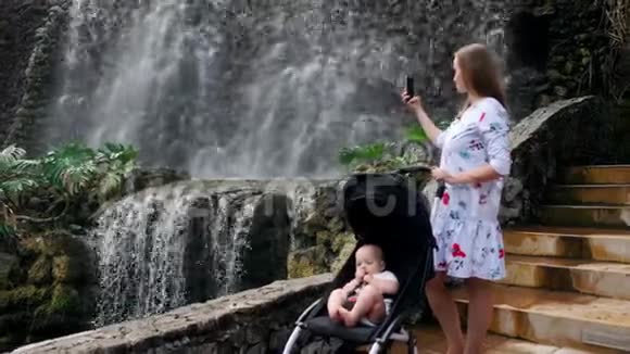 一位年轻的母亲带着她的孩子在瀑布附近散步在她的智能手机上拍照用于社交网络和视频的预览图