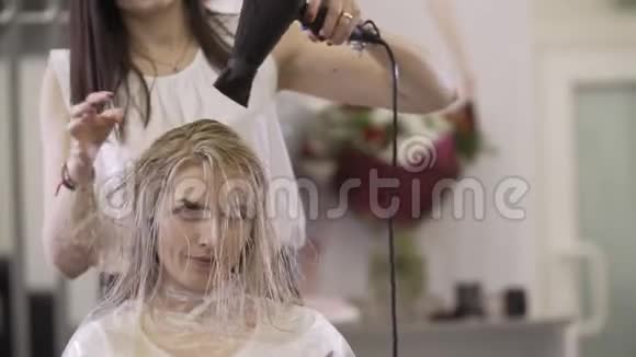 美容院配有吹风机的女造型师一位年轻的金发女郎视频的预览图