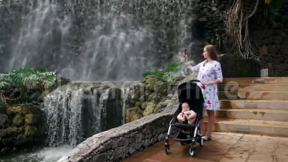 一位年轻的母亲带着她的孩子在瀑布附近散步在她的智能手机上拍照用于社交网络和视频的预览图