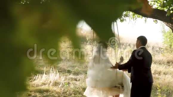 新娘穿着白色连衣裙新郎在公园里荡秋千在橡树的树枝上摇摆视频的预览图