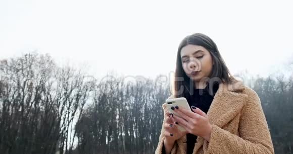 在森林附近的秋天公园年轻迷人的女人微笑着看着智能手机的肖像视频的预览图