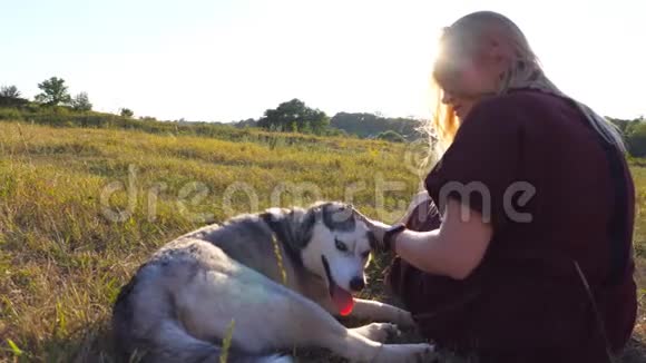 多莉拍摄了一个美丽的女孩坐在草地上在日落时抚摸她的西伯利亚哈士奇狗年轻女子视频的预览图