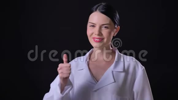自信的年轻女科学家穿着白大褂竖起大拇指对着镜头微笑在黑色背景被孤立视频的预览图