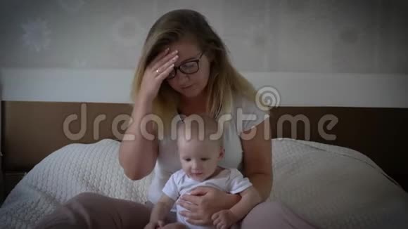 疲惫的心烦意乱的女人头痛手牵着婴儿坐在家里的床上视频的预览图