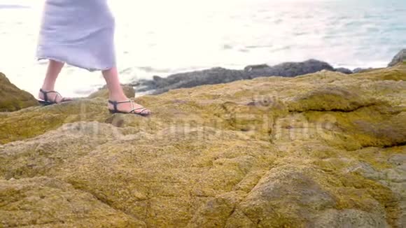 特写镜头女人穿着凉鞋腿和一条长长的灰色裙子在低潮的岩石海岸上视频的预览图