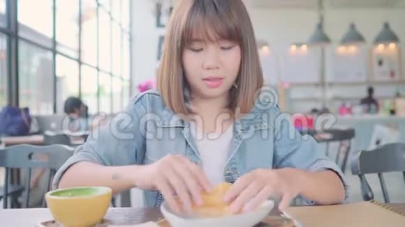 商业自由职业者亚洲妇女吃面包喝温暖的咖啡而坐在桌子上的咖啡馆视频的预览图