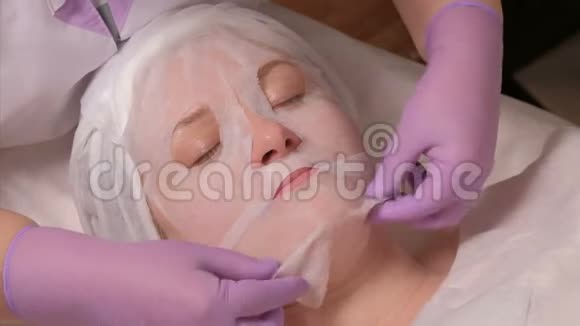 专业美容师戴着紫丁香手套抚平中年女性脸上的面具皮肤的程序视频的预览图