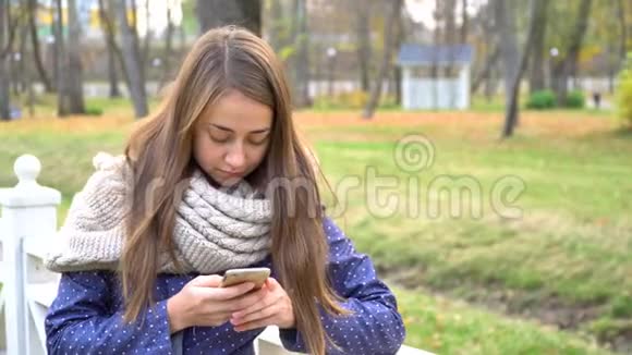 白人妇女戴着针织围巾使用智能手机在秋天公园散步时打字视频4k视频的预览图