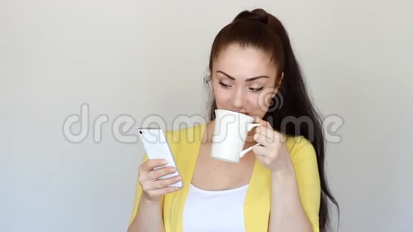 年轻女性从杯子里喝咖啡使用互联网应用程序在智能手机屏幕上看手机应用程序视频的预览图
