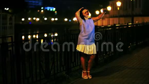 年轻的微笑女人在码头和夜空的背景下在手机上自拍视频的预览图