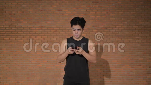 年轻快乐英俊的亚洲人用电话对着砖墙思考视频的预览图