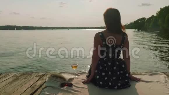 穿着衣服的女人坐在湖边的老码头上试着喝酒但笑着把一杯酒放回水面视频的预览图