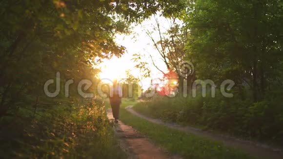 红发女人穿着裤子穿着白色衬衫走着夕阳西下的乡间小路上有美丽的傍晚阳光绿色的阳光视频的预览图