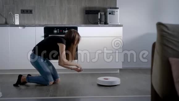 布鲁内特女孩在厨房里玩吸尘器机器人吸尘地板视频的预览图