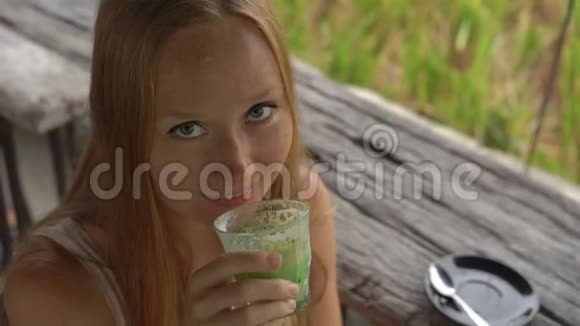 一位年轻女子在一家热带咖啡馆喝绿茶抹茶拿铁时的慢镜头观看稻田视频的预览图