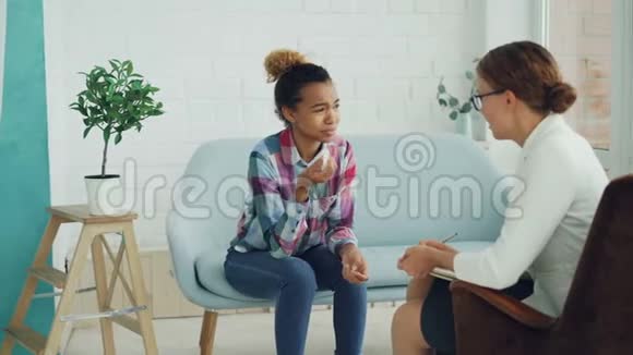 不高兴的混血女孩正在和心理学家交谈坐在沙发上哭泣而医生则抱着她听着视频的预览图