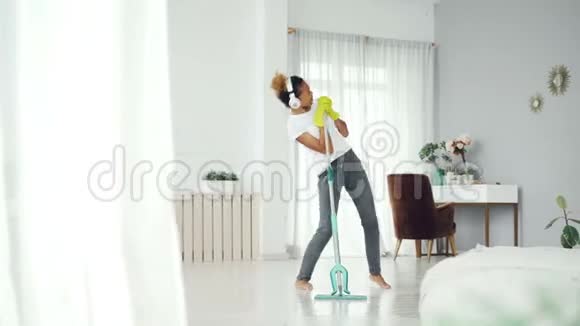 有趣的非洲裔美国青少年正在打扫房子做家务用拖把清理地板在里面听音乐视频的预览图