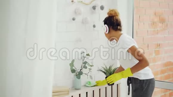 开朗的女佣非裔美国女孩正在做家务用布擦灰尘戴手套年轻的女人在听视频的预览图