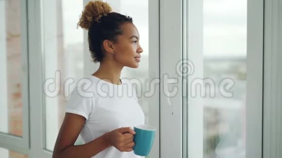 快乐的混血女人正望着窗外喝着咖啡站在大窗户附近享受着城市的乐趣视频的预览图