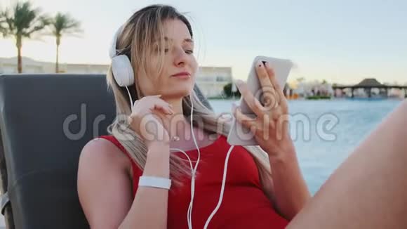 穿着红色比基尼带着白色耳机听智能手机音乐的年轻迷人的微笑少女的肖像视频的预览图