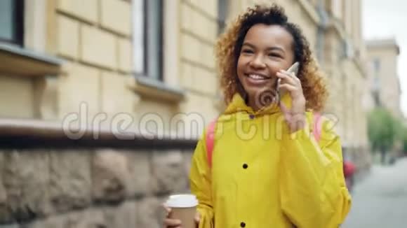 快乐的混血少女正在用智能手机说话拿着咖啡走在美丽的大街上视频的预览图