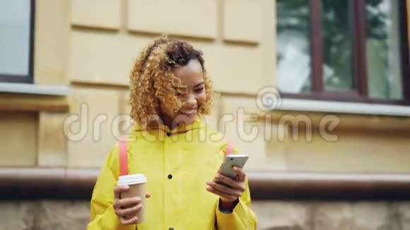 漂亮的非洲裔美国女孩看着智能手机屏幕微笑着站在户外穿着明亮的衣服触摸它视频的预览图