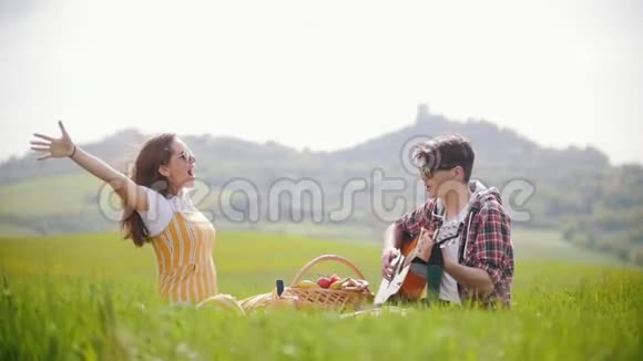 一对年轻夫妇穿着鲜艳的衣服坐在明亮的绿色草地上一个男人弹吉他而他的幸福女友视频的预览图