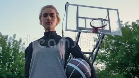 年轻漂亮的女子篮球运动员拿着球看着镜头站在公园里低角度视频的预览图