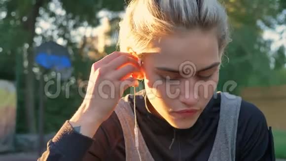 美丽的金发女郎戴着耳机戴着刺鼻的鼻孔日落时坐在公园里向前看视频的预览图