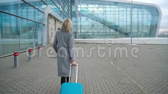 金发女孩在机场航站楼附近滚动一个手提箱从后面看慢动作视频的预览图