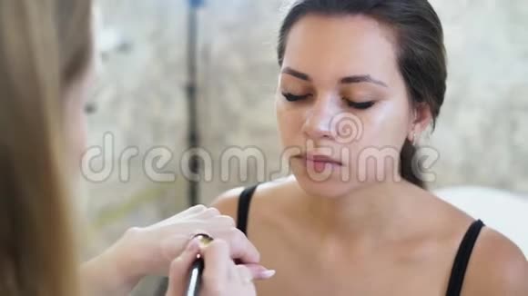 一位年轻的金发碧眼的化妆师用一个遮瑕膏在她脸上画一个年轻的黑发女孩视频的预览图