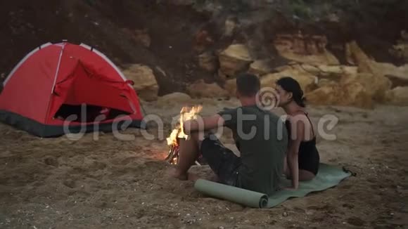 一对无忧无虑的年轻夫妇晚上坐在空荡荡的海滩上的篝火旁女孩在视频的预览图