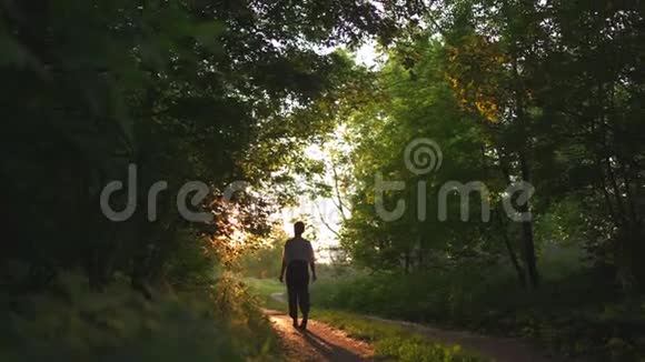 红发女人穿着裤子穿着白色衬衫走着夕阳西下的乡间小路上有美丽的傍晚阳光绿色的阳光视频的预览图
