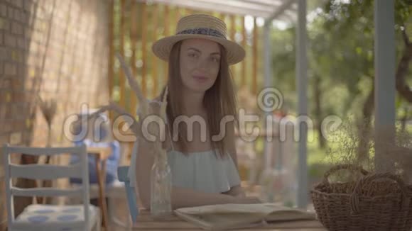 美丽的年轻女孩戴着草帽和白色连衣裙坐在户外的小木桌上看书农村视频的预览图