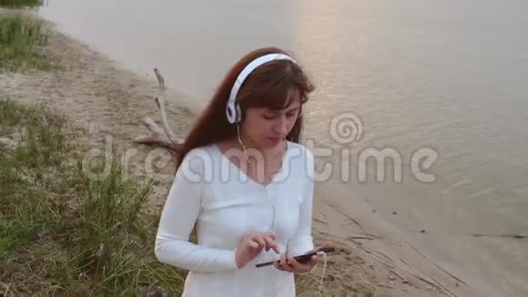 戴着耳机的女孩走在水旁边的沙滩上听着平板电脑上的音乐视频的预览图
