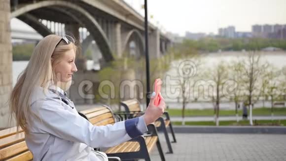 年轻漂亮的金发女郎用智能手机自拍一位年轻女子沿着河堤散步视频的预览图