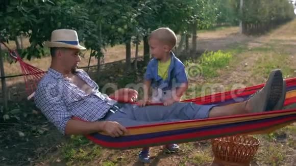 户外娱乐活动孩子在苹果花园的树间的吊床上摇着父亲视频的预览图