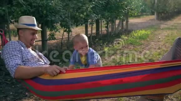 快乐的时刻孩子在苹果园水果篮附近的吊床上摇着父亲视频的预览图