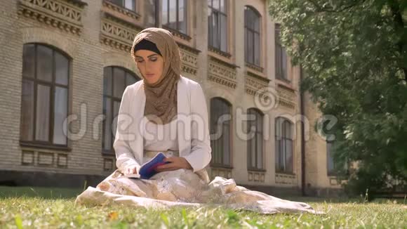 美丽的穆斯林妇女戴着头巾坐在公园的草地上看着书环顾四周在背景背景上视频的预览图