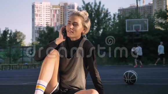 快乐的女篮球运动员打电话微笑着坐在露天的球场上男人在后台玩视频的预览图