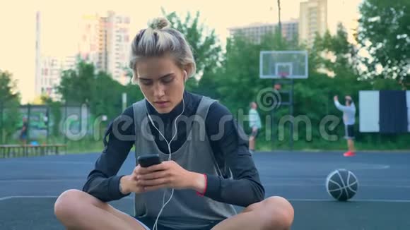 漂亮的年轻女子坐在篮球场上打电话戴着耳机在公园里停着摩天大楼视频的预览图